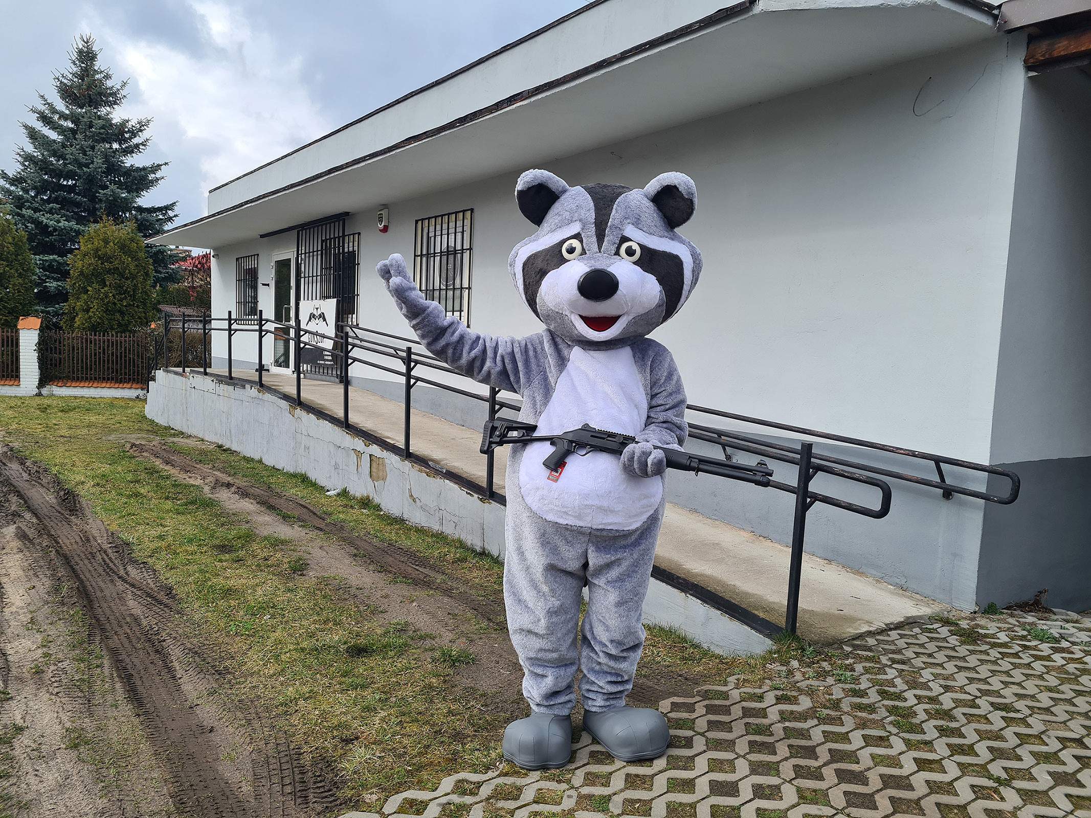 Na fotografii widnieje kostium reklamowy w postaci szopa pracza pozujący do zdjęcia przed siedzibą firmy GunSzop